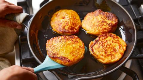Four golden potato cakes sit in a frying pan. Someone is flipping one with a spatula.