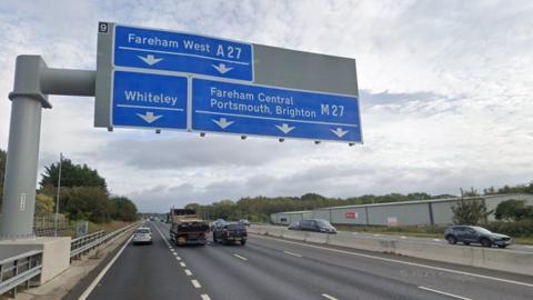 Blue and grey motorway signs above four lanes of motorway.