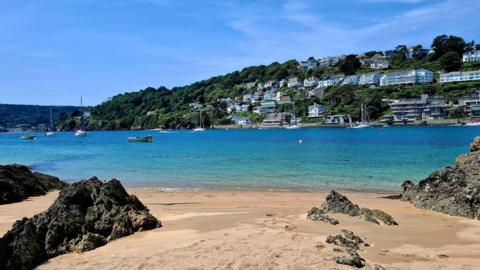 View of sandy beach with large brown rocks, boats in turquoise sea, houses on coastline, under a blue sky
