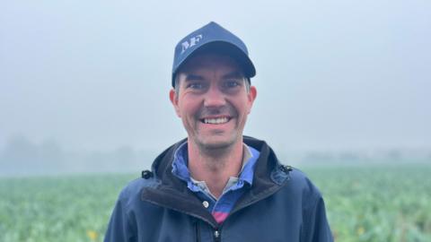 Phil Le Maistre is a man with short hair. He is wearing a navy baseball cap with a blue and pink collared shirt, underneath a navy waterproof. The background is an out-of-focus misty field.