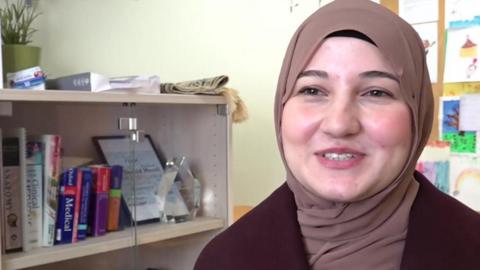 A woman in a pale brown head scarf and maroon top looks at the camera. She is sitting in an office. To her right is a glass-fronted cabinet, with books inside and a plant on top of it. The wall is pale yellow in colour.