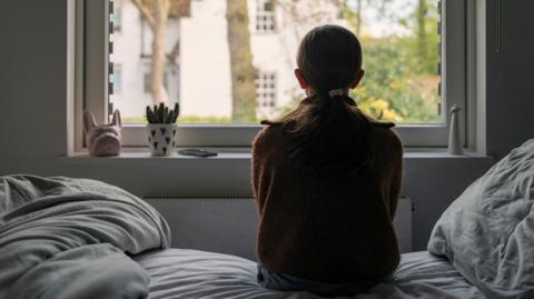 Pictured from behind, a girl looks out of a bedroom window