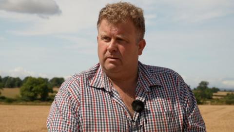 William Maughan, a middle aged man with blonde, curly hair. He is stood in the middle of a harvested wheat field, which stretches out into a backdrop of rolling, beige hills under a light blue sky.
