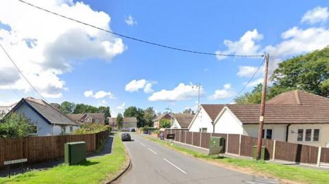 An "Oak Avenue" signpost can be seen to left on a grass verge. There is a road down the middle with homes on both sides and at the end. Wooden fences line each side.