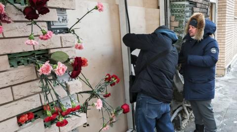 Municipal workers in puffer jackets inspect a blown out door that has been boarded up outside the apartment building where Kirillov died. Plastic flowers are placed in exposed brickwork