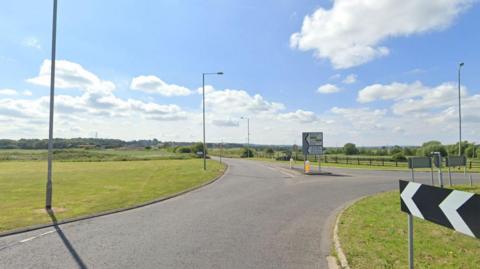A view of one of the roundabouts and B-roads that already exist that will have the development around it. Empty roundabout with round going straight off ahead on a sunny day with green fields around