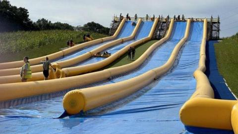 A long, sloping, blue plastic mat on a grassy hillside with lanes divided by inflated yellow walls.