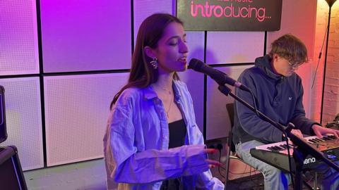 Nina Arya sits on a stool in a studio and is singing into a microphone. She wears an over-sized blue shirt. A man with glasses plays a keyboard. There is a purple light glowing and the BBC Introducing sign is on a wall.