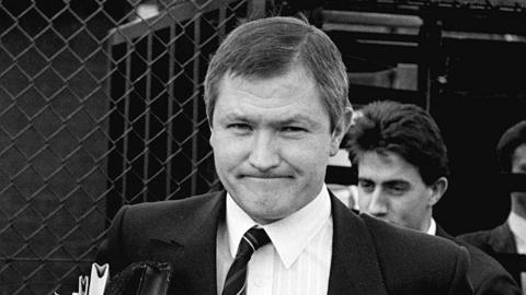 A black and white still of Pat Finucane, who is wearing a suit jacket, a shirt and striped tie. He is walking out of a court building while holding a folder and files.