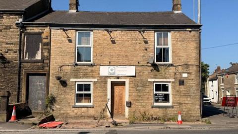 Image shows a dilapidated pub on the end of a street. It is built of stone and its front door is boarded over. 