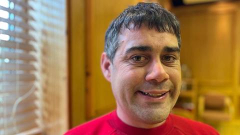 Tom has short, dark hair and is wearing a red t-shirt. He is in a room used by the organisation People First and is smiling at the camera.