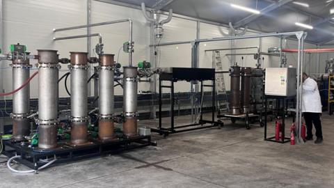 A large mechanical device with tall stacks connected to other equipment installed in a warehouse being operated by a man in a white coat
