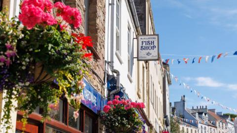 A view of a quaint street, showing rows of shops complete with hanging baskets and colorful bunting.