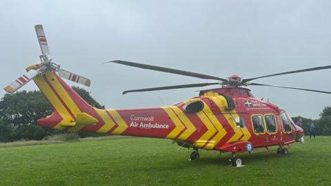 A picture of a Cornwall air ambulance parked at a field. It is red and orange in coloured and is parked in front of a goal post. 
