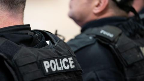 The back of two police officers in full uniform. Both wear bullet-proof vests with the word police written on the back. 