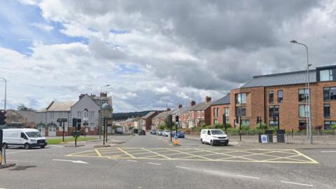 View of a crossroad junction, showing the central yellow hatched area. There are traffic lights with stationary traffic including two white vans and a number of cars. To the left of the road directly ahead is a large building with white plaster upper storey and brick ground floor, and to the left a two-storey brick building with a number of windows. Terraced residential properties line the road stretching into the distance.