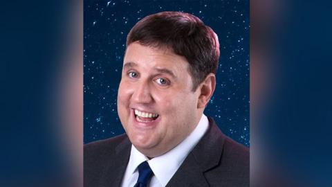 Close up of comedian Peter Kay with brown hair wearing a dark grey suit and navy tie. He is standing in front of a background of navy blue and glitter. He is smiling.