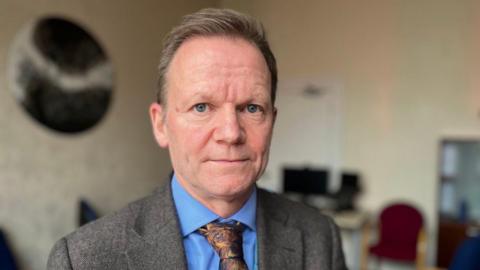 Joe Teape is wearing a blue shirt, a brown patterned tie and a brown suit jacket and is in an office with the background blurred