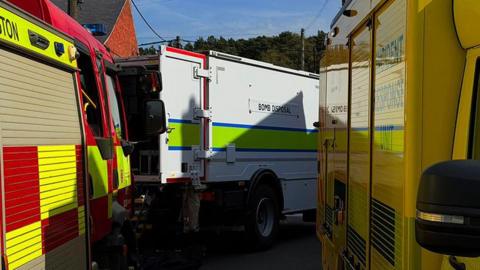 A red and yellow fire engine, white bomb disposal van and yellow incident response unit van all parked extremely close to each other. The fire engine is on the left and incident response on the right, with the bomb disposal van parked an an angle in the middle. The back doors of the bomb disposal van are open. The top of a red brick house is visible above the vans on the left. There are telephone wires above them and trees peaking out beyond the bomb disposal van. 