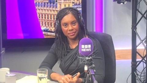 Kemi Badenoch in a BBC Radio studio, sitting in front of a purple microphone that says BBC News. She is wearing a black top, and behind her is a screen shwowing an image of the Houses of Parliament 
