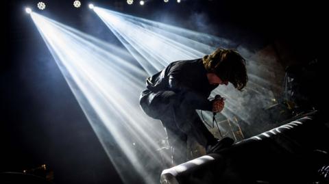 A man crouches on stage. He is holding a microphone and wearing all black. He has long, dark hair. His upper-body is parallel to the ground, and he is illuminated by two spotlights from above. 
