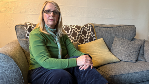 A woman wearing a green top and jeans sits on grey sofa. Her hands are clasped together and lying across her left knee. A number of cushions can be seen on the sofa behind her.