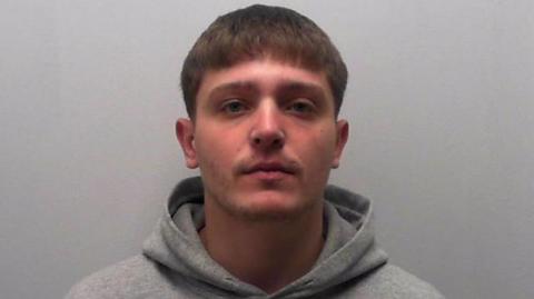 Image of a young man in grey hoodie at a police station. He has short brown hair. He is standing in front of a grey background.