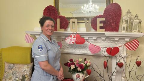 A woman in a carer's uniform standing beside a fireplace smiling at the camera. The fireplace is decorated with hearts and flowers for Valentine's Day