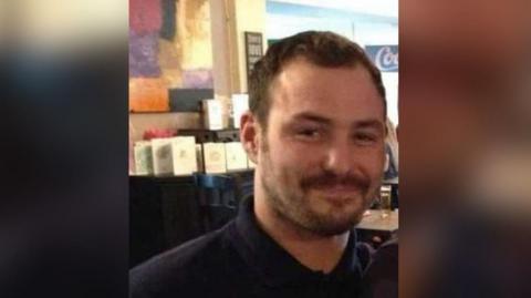 Man who has short dark hair with a short dark beard and smiling, he is wearing a dark polo top with a collar and is sitting in a bar with tables behind him.