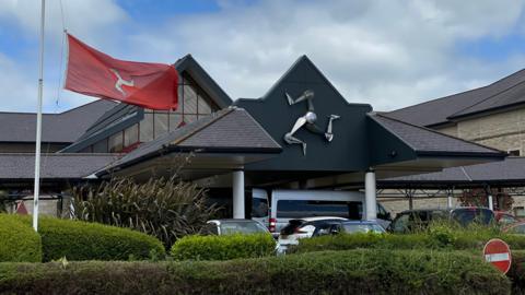 The exterior of Noble's Hospital. The pointed roof of the front canopy features a silver three legs of Man sculpture. There are cars parked outside and a Manx flag can be seen flying from a flag pole.