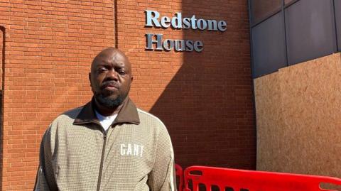 A man with short dark hair and a beard, wearing a beige zip-up jacket. He is standing in front of a brick-built block of flats with several storeys, with a glass entrance porch and "Redstone House" in silver lettering on a brick wall. There is wooden boarding around the entrance porch, and a low red temporary fence in front of the entrance.