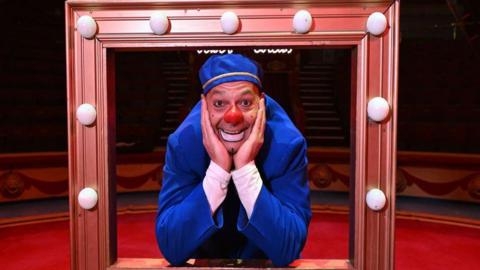 A man is pictured in a circus ring dressed as a clown, with a red nose and white face paint around his mouth. He is wearing a blue jacket and hat with white sleeves seen under his jacket. He is smiling while posing with his elbows resting on a dressing room mirror frame, with light bulbs around it.