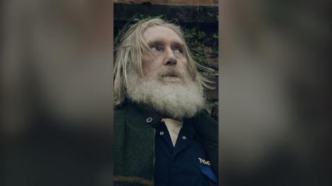 A man with long grey hair and a beard stands by a wall looking to the side.