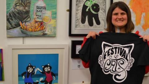 A woman holding a black and white t-shirt with the words Festival of Cats. She is standing against a series of artwork that feature cats.