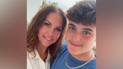 A woman wearing a white blouse looking at the camera smiling, alongside her teenage son who is also smiling and wearing a blue T-shirt and silver necklace in the selfie picture.