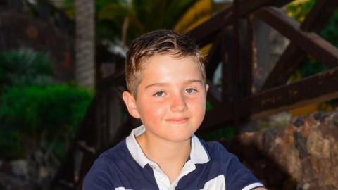 Ronnie smiles and looks directly at the camera as he is photographed outside, wearing a blue and white polo shirt.