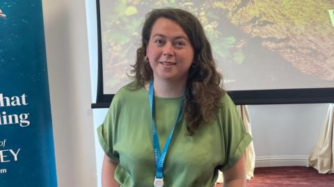 Jo Ferguson is wearing a green blouse and a turquoise lanyard. She has long dark hair and is smiling at the camera. She is standing in front of a giant screen which has a photos showing shrubs and grass. To her right is part of a banner.