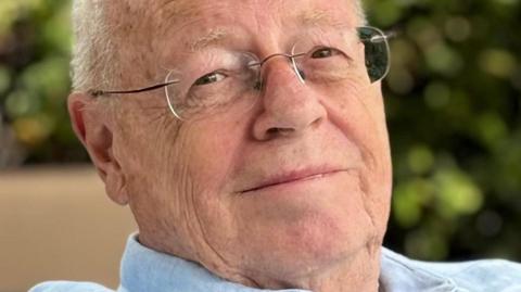 Mark Talbot, a man in his 70s, wearing a blue shirt and glasses is sitting in an armchair and smiling. He has short white hair and there is greenery behind him.