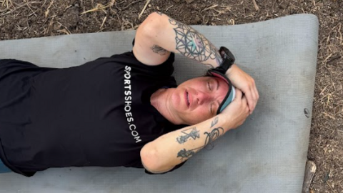 Hannah lies on yoga mat. She wears a black t-shirt and light blue sports cap and has tattoos on both arms. Her eyes are shut as though she is very tired and she is lying outside on rough ground with grass or straw.
