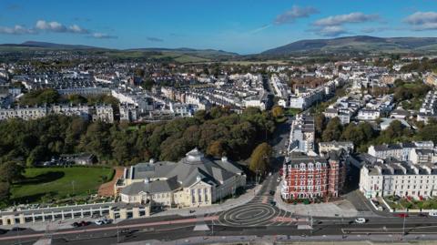 An aerial view of the road along Douglas promenade, you can see the roundell in the middle and the Villa Marina on the left of the picture, there are houses in the background and hills.