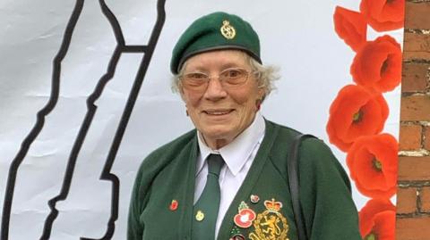 A smiling Sandy Cawley wears a green beret, tie and cardigan with army badges and insignia. She wears glasses and a white shirt, with a black handbag over one shoulder. 