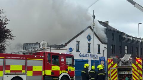 Two red and yellow fire engines parked in front of Galaxy Blinds which is a white and blue building with a pointed front. Grey smoke covered the entirety of the roof and is billowing upwards and to the right. Four firefighters are standing in front with yellow helmets on. There is a row of grey terraced buildings to the right.