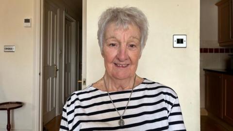 A woman with short grey hair smiling at the camera. She is wearing a black and white striped top. She also wears a silver necklace with an anchor pendant on it 