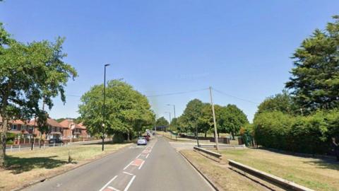 A suburban road flanked by grass verges, hedgerows and trees. Telegraph poles, lamp-posts and other street furniture can be seen. It is a sunny day.