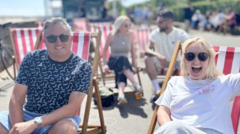 A man and woman sitting on deck chairs in the sunshine. both of them are smiling, with the man wearing a blue t shirt and denim shorts and the woman has a white T shirt on. There are people behind them also sitting on stripy deck chairs