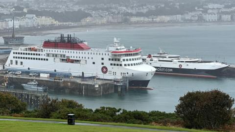 The Manxman and Manannan ferries moored in Douglas Harbour. The are painted in the colours of the Steam Packet Company of red, white and black. The sweep of Douglas Bay the buildings on Douglas Promenade provide a backdrop to the vessels.
