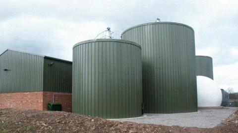 Three large green metal tanks in front of a building with a green metal roof and red bricks.