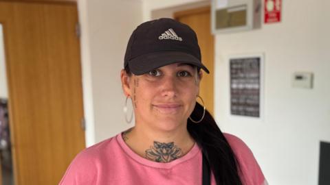 A head and shoulders image of Becky Mather. She is wearing a baseball cap and a pink jumper. She is looking into the camera. She has a large floral tattoo on her throat, and smaller tattoos down both sides of her face