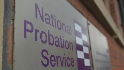 A sign with the logo of the National Probation Service written on it, attached to a red brick wall.