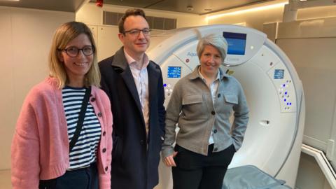 Dr Claudia Wegner, wearing a stripy top and pink cardigan, Dr Henry Steer, wearing a light dark jacket and white shirt and Dr Anna Bibby wearing a light shirt and grey jacket, all standing by a CT scanner.